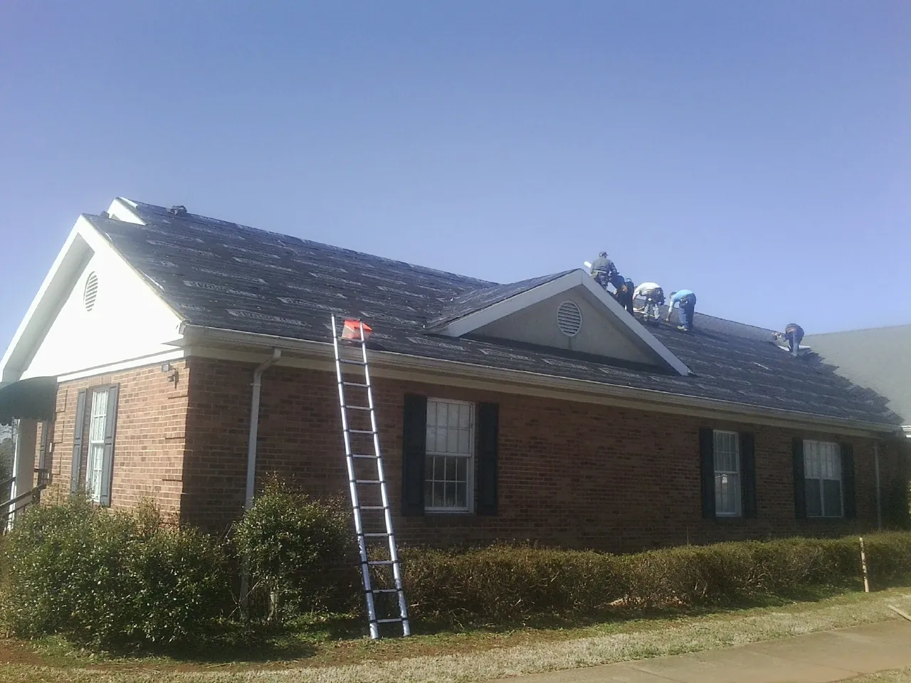 Crew tearing shingles off a brick ranch roof with a ladder set — Metal Roof Repair work by our crew in Masonville
