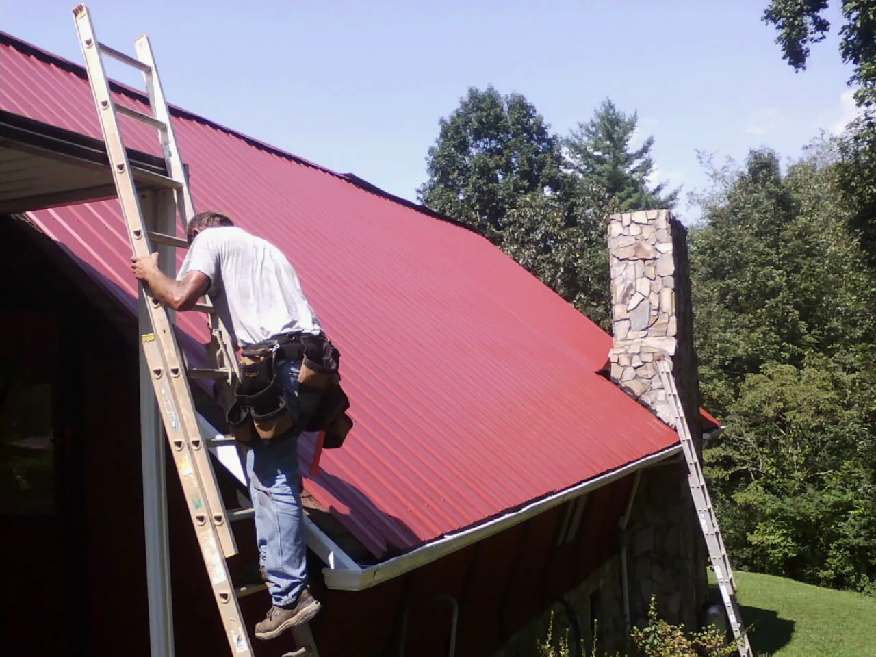 Roofer on a ladder beside a red metal roof and stone chimney — Soffit & Fascia Repair by Roofing Brothers in Masonville