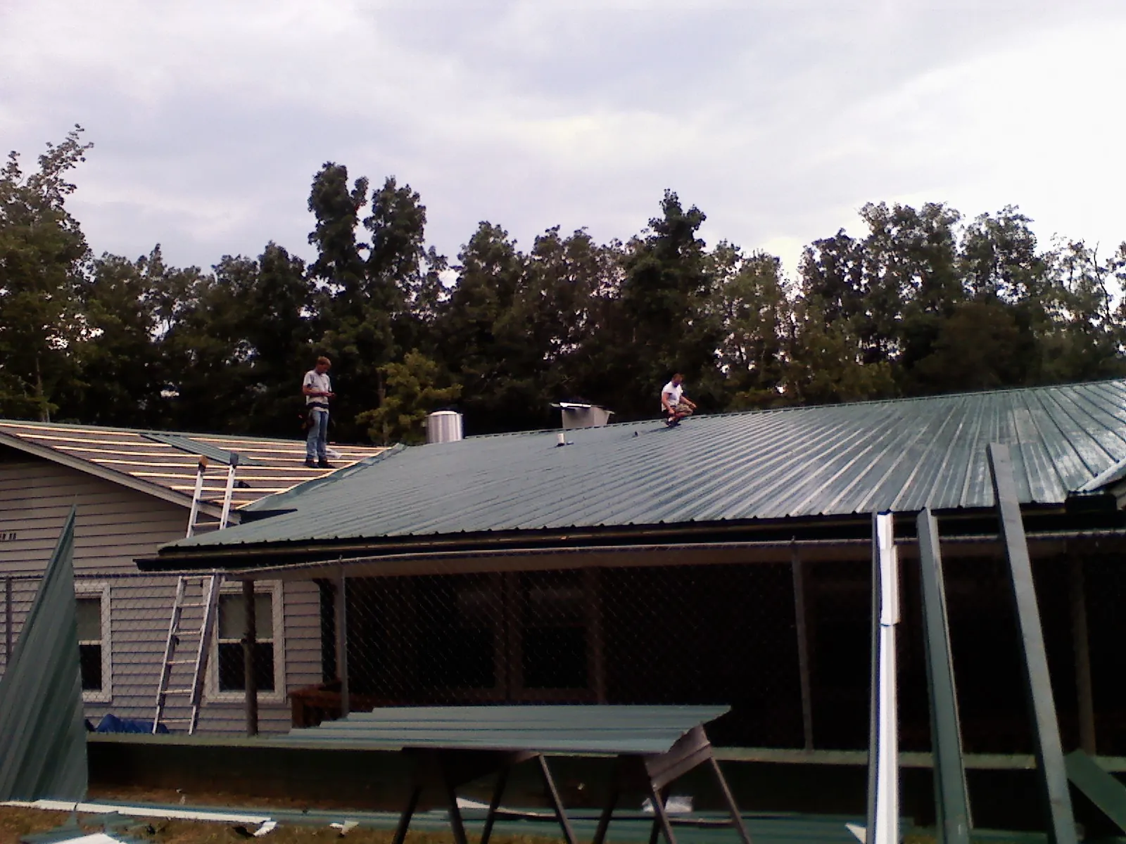 Two roofers installing green metal panels above a covered porch in Masonville